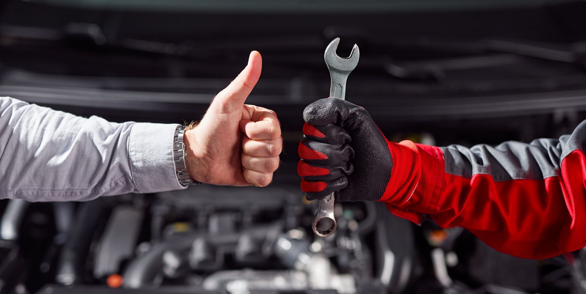 Hands of a customer & mechanic. Customer gives thumbs up. Mechanic holds wrench, wears red & gray work clothes, over engine.