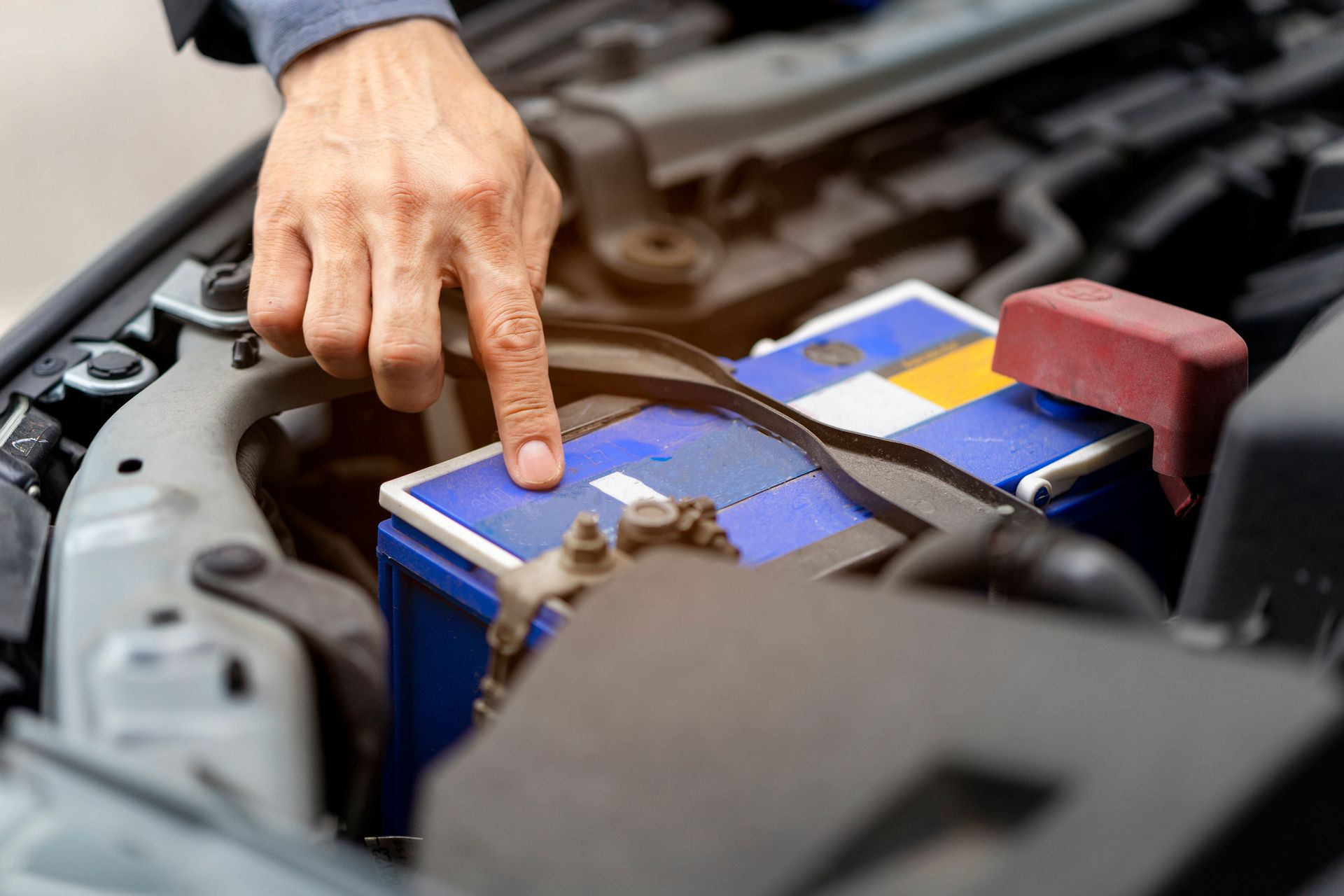 Person's hand pointing at a car battery in engine compartment; blue and yellow battery, black belt, and other components.