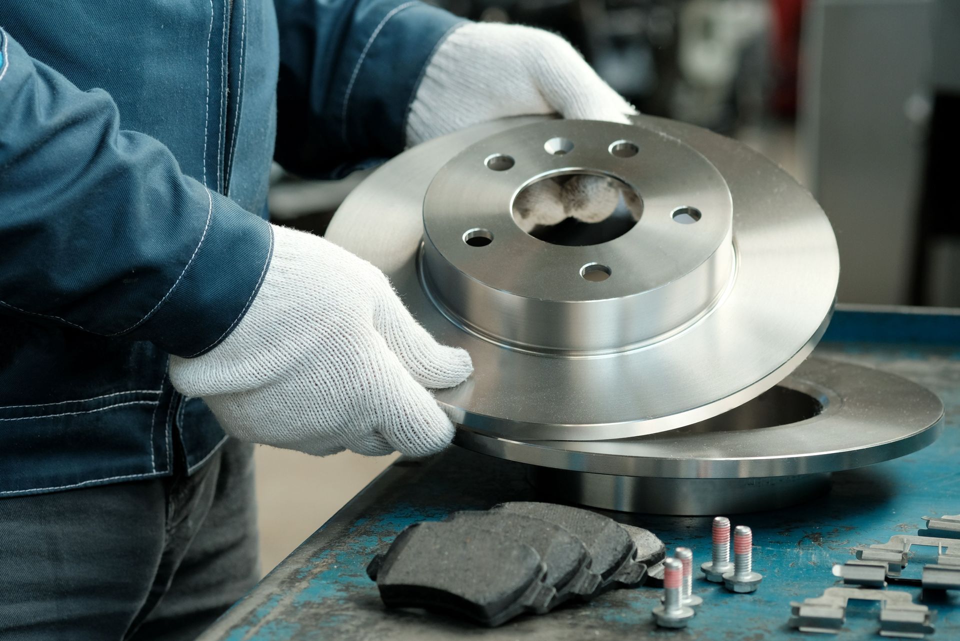 Mechanic wearing gloves holding a new brake rotor, with brake pads and bolts on a table.