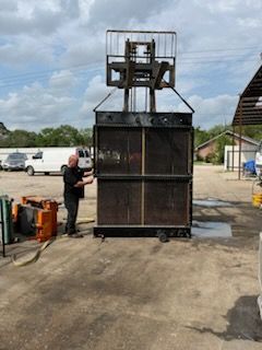 Man standing next to a large black metal compactor, with a forklift at the top, outdoors.