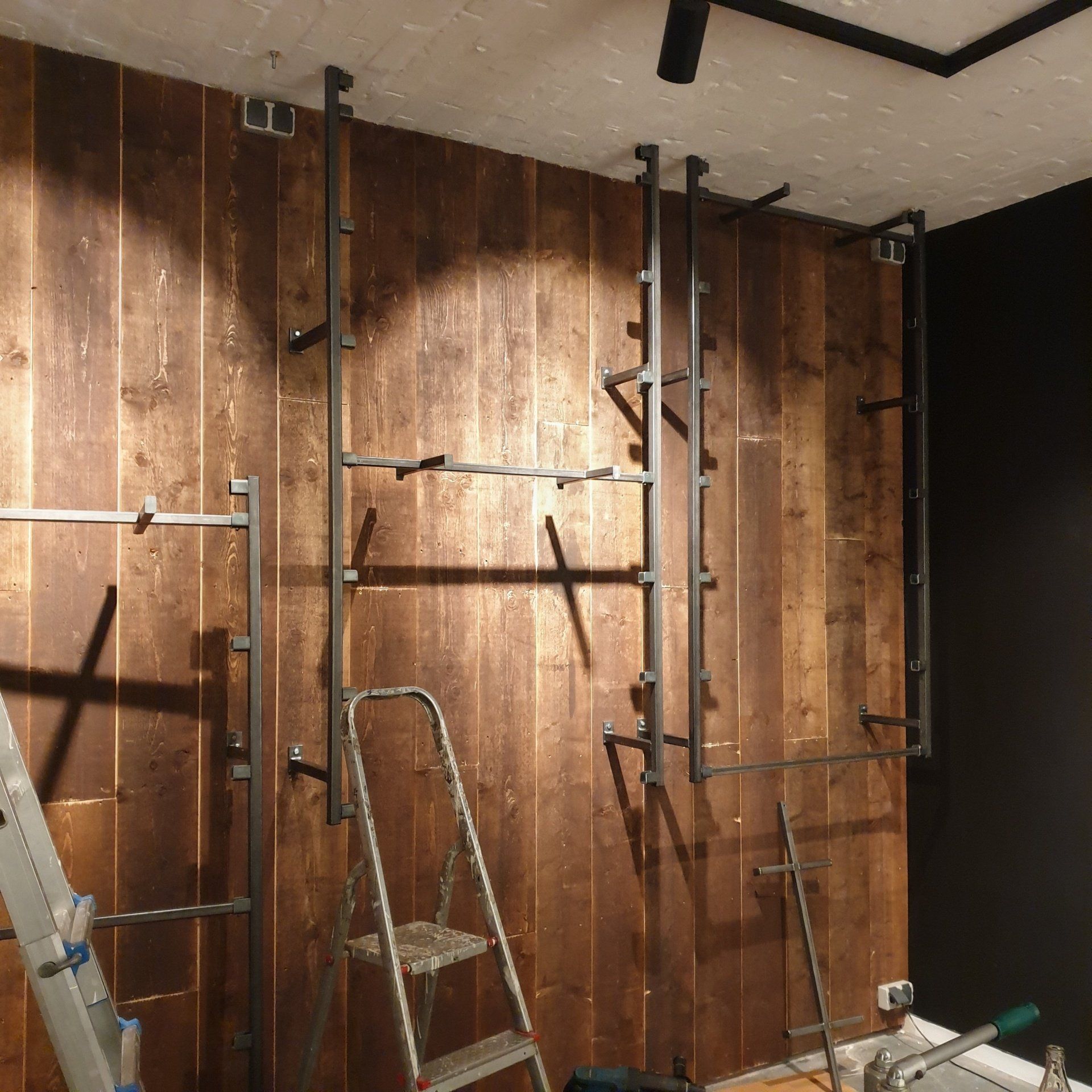 Metalen stellingframes gemonteerd op een rustieke houten plankenwand, met een trapladder ernaast in een bouwomgeving.