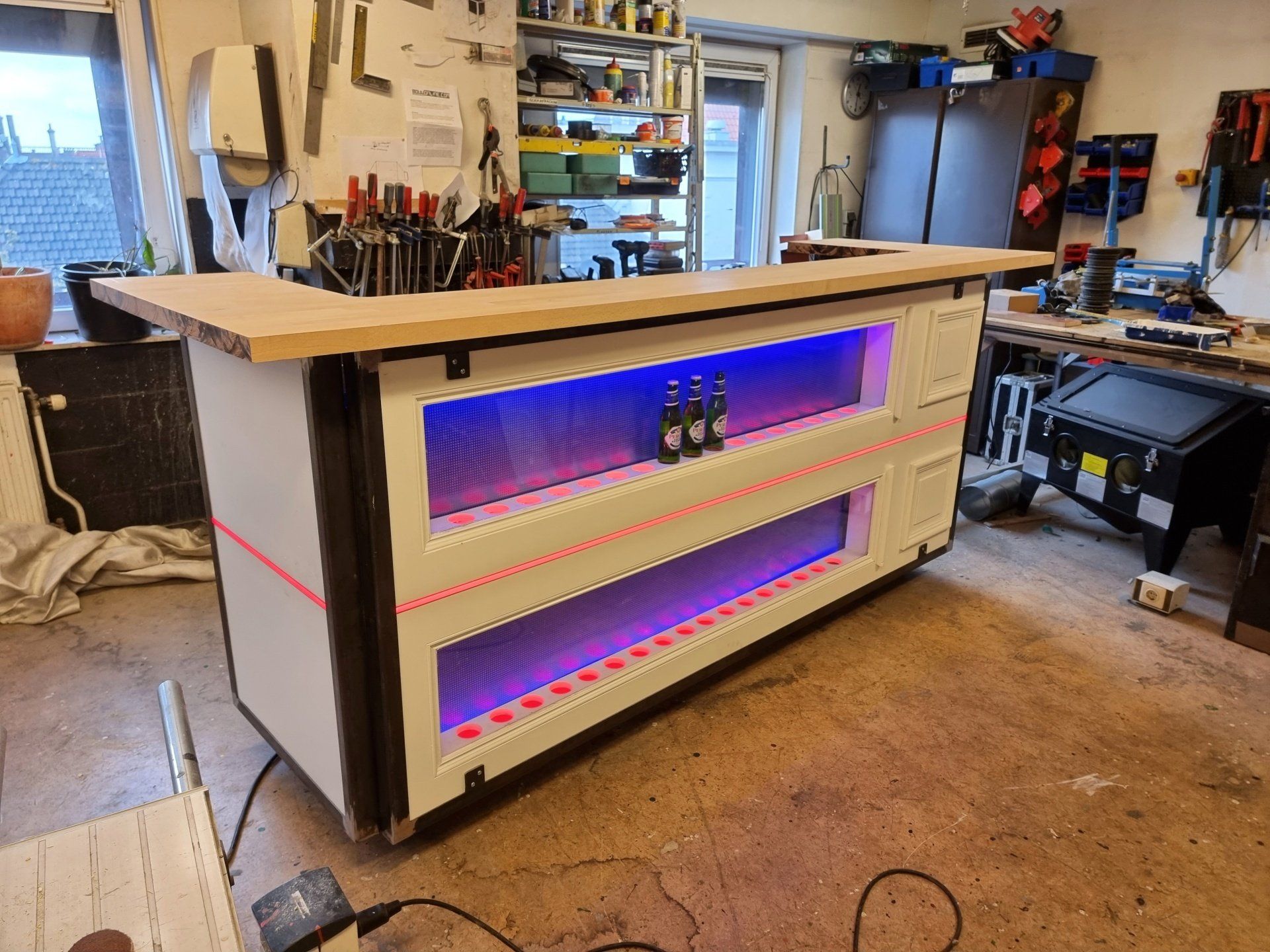 Een op maat gemaakte thuisbar, gemaakt van witte panelen met twee paars oplichtende planken, geplaatst in een rommelige houtbewerkingswerkplaats.