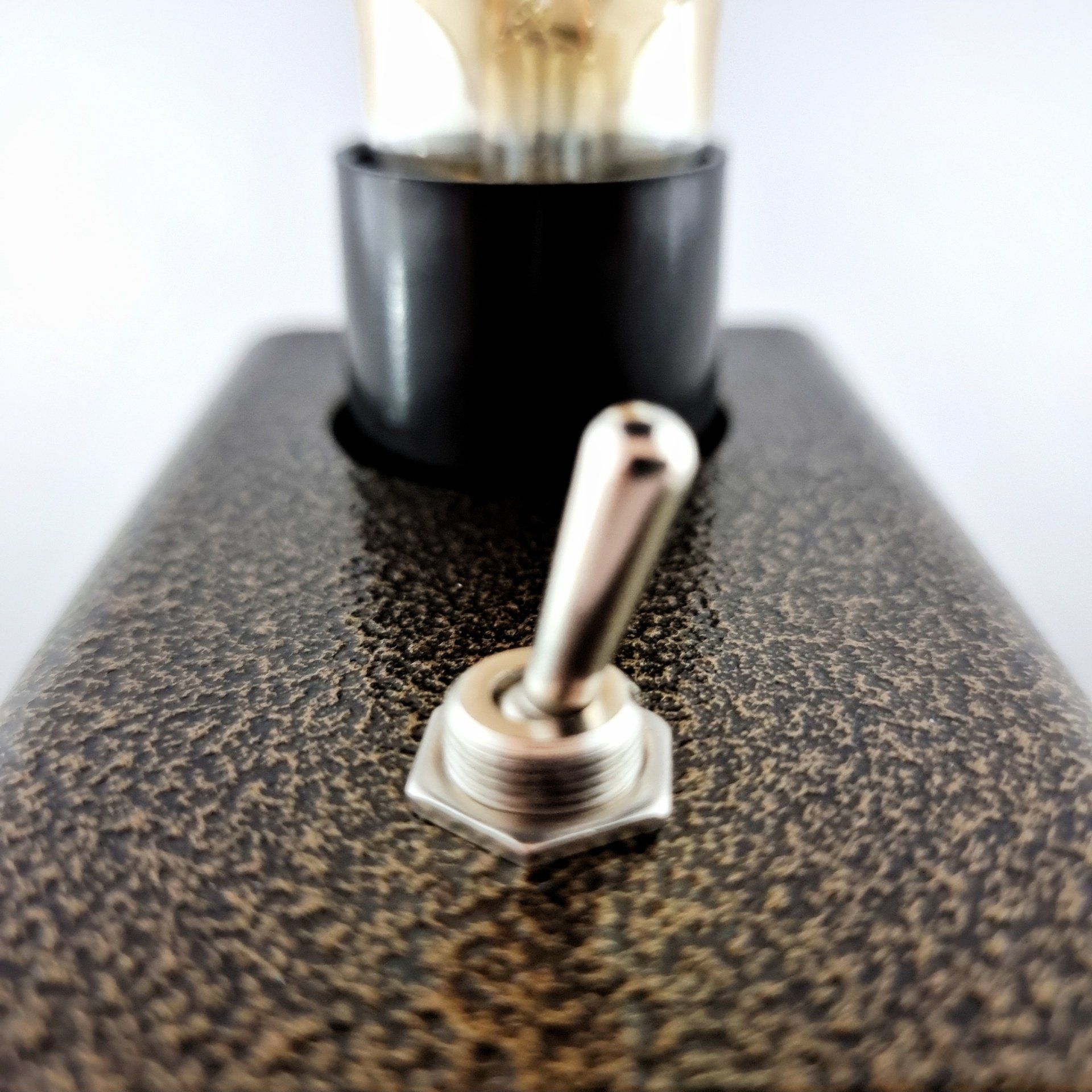 Een close-up van een metalen tuimelschakelaar op een gestructureerde, bronskleurige lampvoet, met daarboven de voet van een gloeilamp zichtbaar.