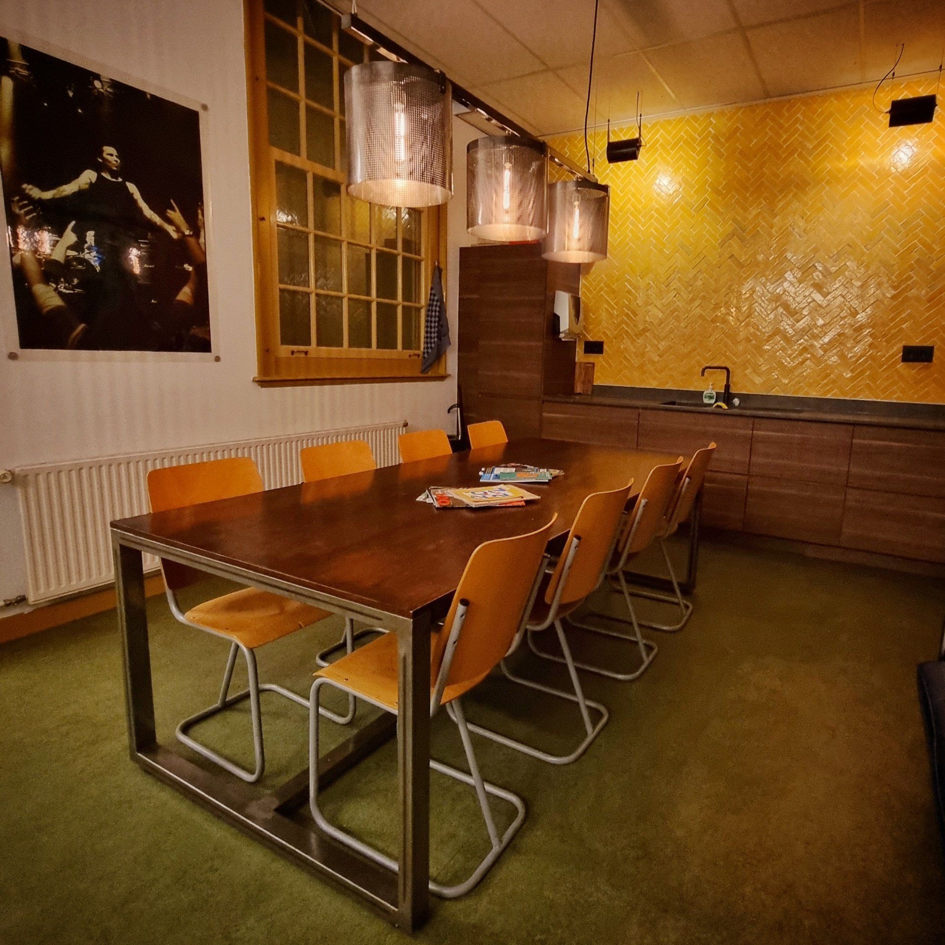 Een eethoek met een lange houten tafel, oranje stoelen, industriële hanglampen en een gele tegelwand in de keuken.
