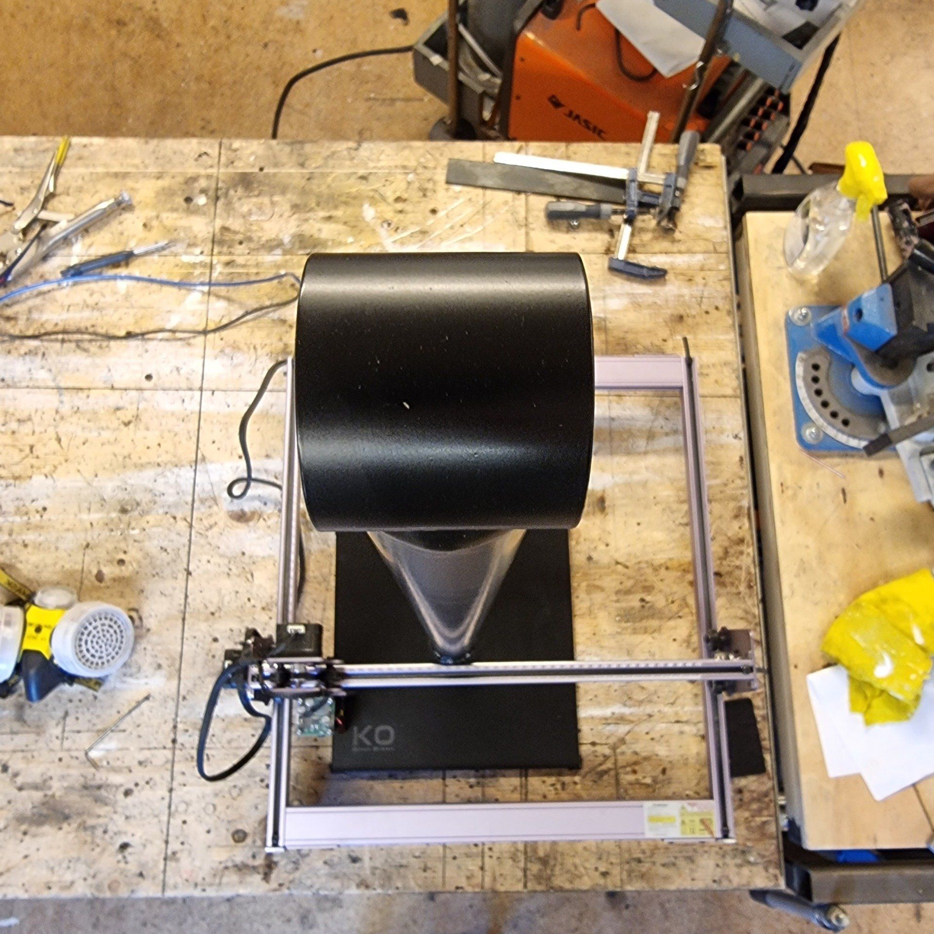 Een tafelmodel lasergraveermachine staat op een houten werkbank, met een zwarte cilindrische afzuigkap boven het werkoppervlak.