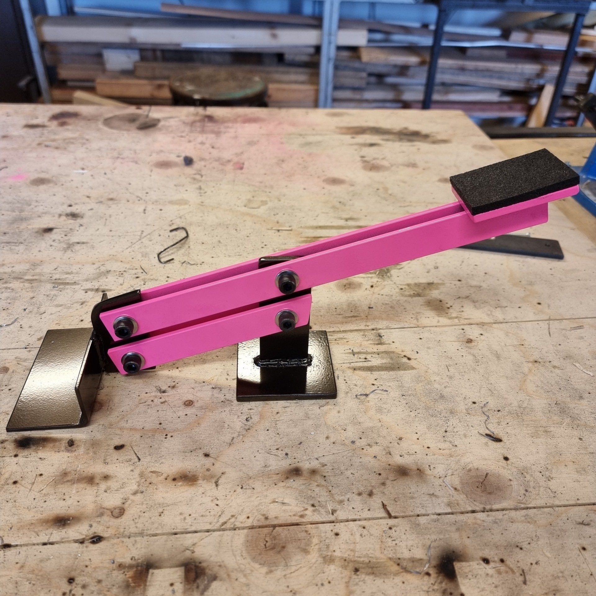 Een roze, scharnierend hefboomgereedschap met een zwarte voet en handgreep, gemonteerd op een werktafel.