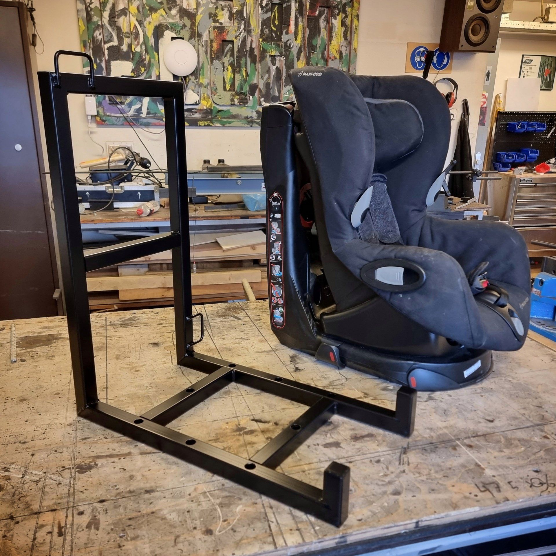 Een zwart metalen frame voor een autostoeltje, naast een zwart kinderautostoeltje op een werkbank.