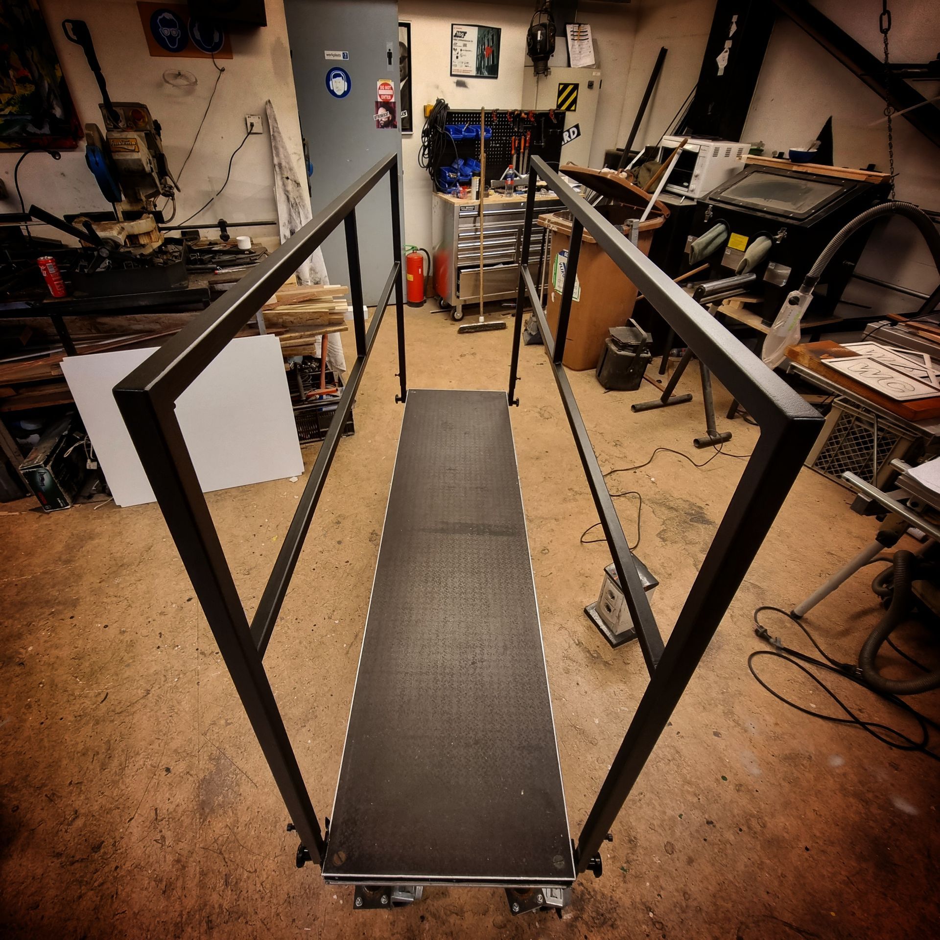 Een metalen loopbrug met zwarte leuningen en een zwart, gestructureerd platform, gelegen in een rommelige werkplaats.
