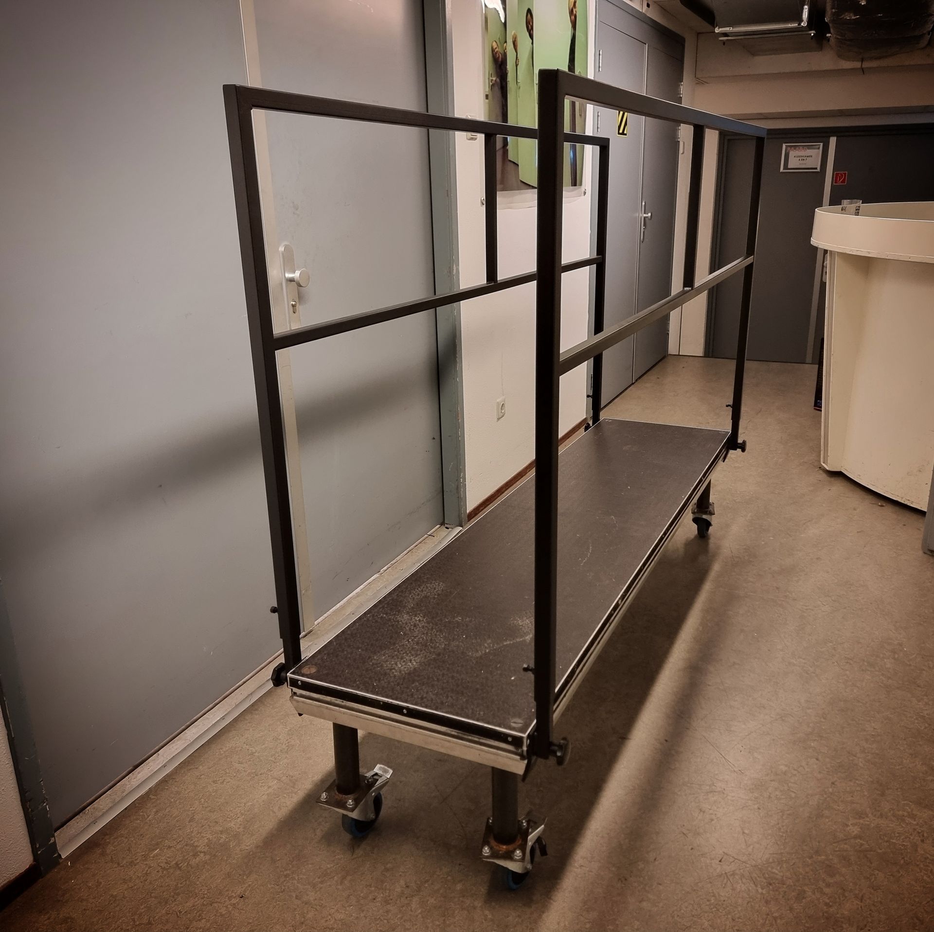 Een verrijdbaar, rechthoekig, zwart podium met metalen veiligheidsleuningen aan beide zijden, geplaatst in een overdekte hal.