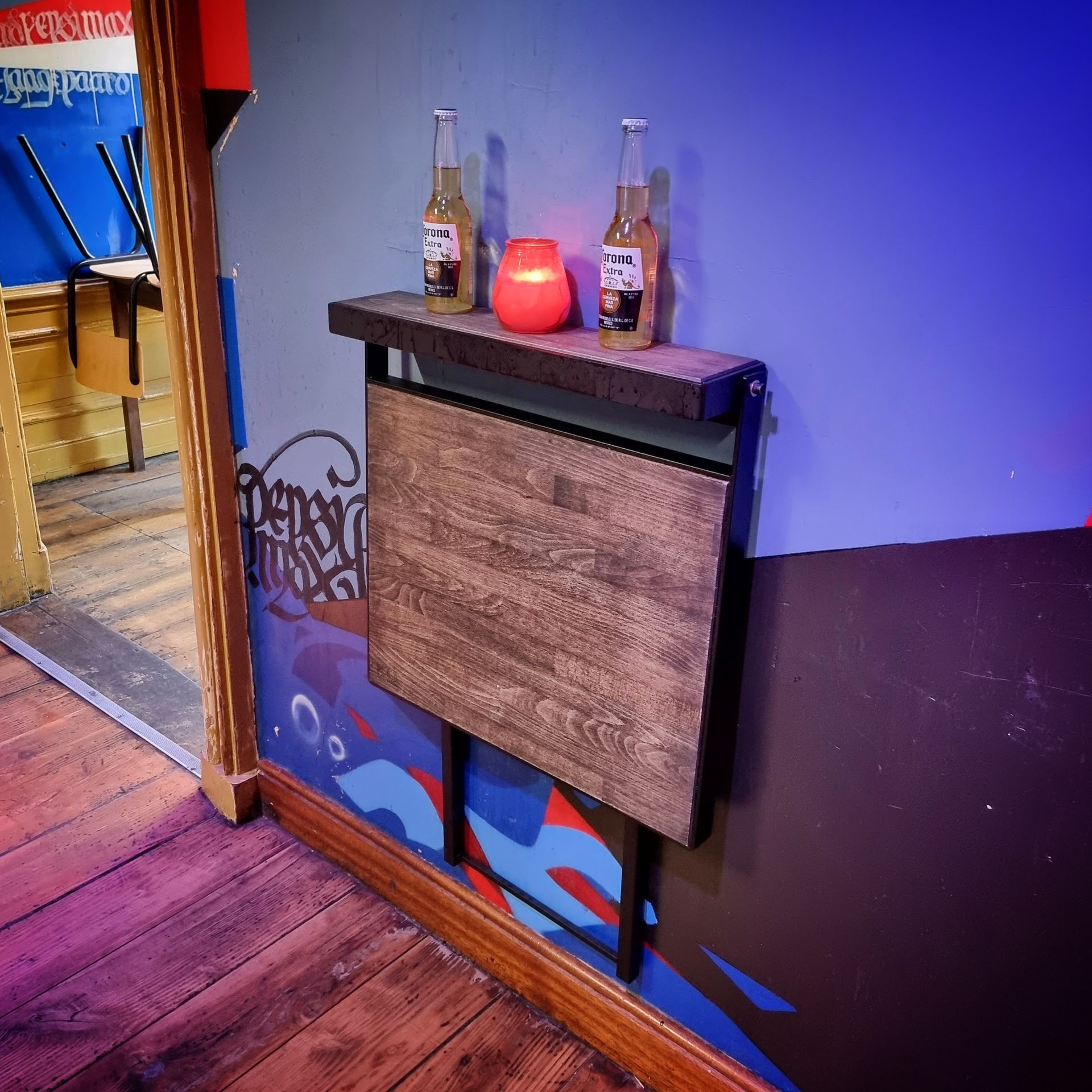 Een houten, aan de muur bevestigde klaptafel in een kamer met blauwe muren, met daarop twee glazen flessen en een gloeiende rode kaars.