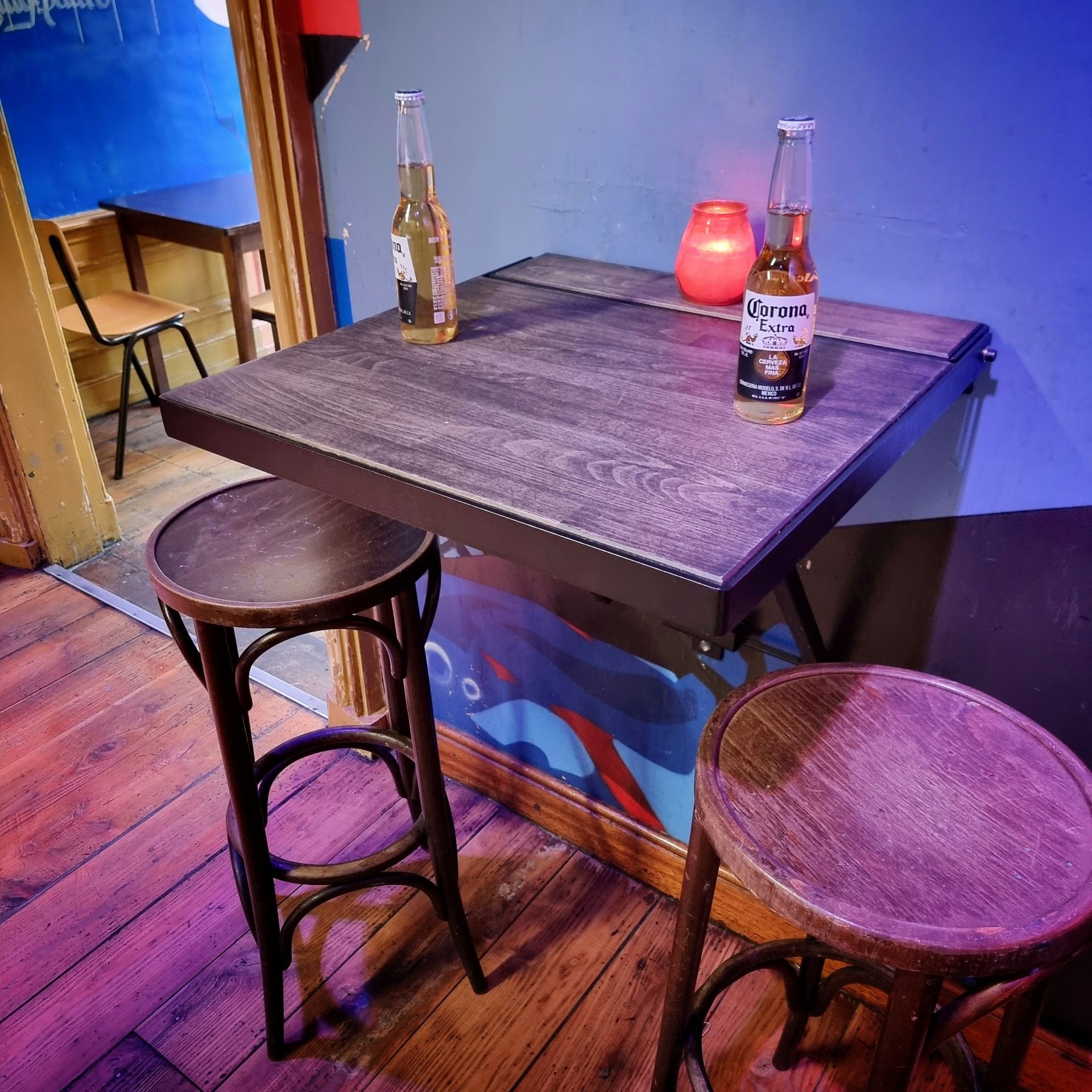 Twee barkrukken staan ​​aan een donkerhouten bartafel met twee flesjes bier en een klein rood lampje tegen een blauwe muur.