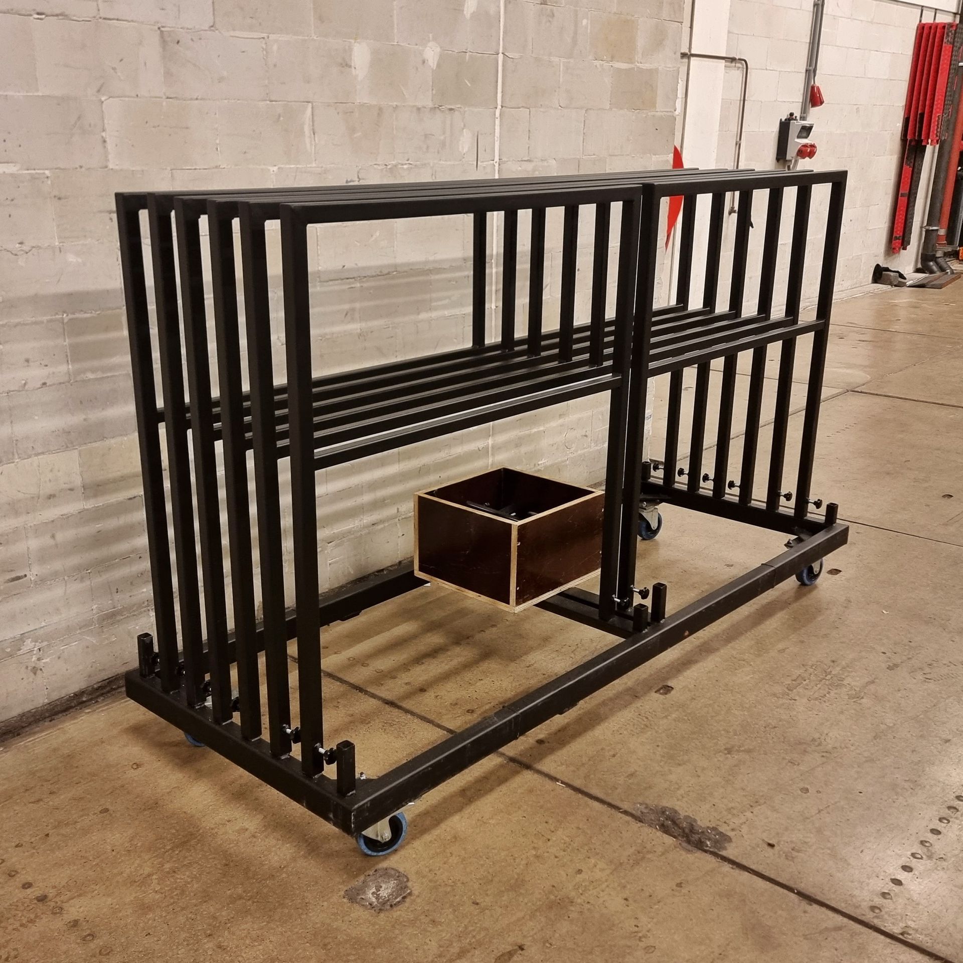 Een zwart metalen rolrek met een systeem van planken staat op een betonnen vloer naast een houten kist.