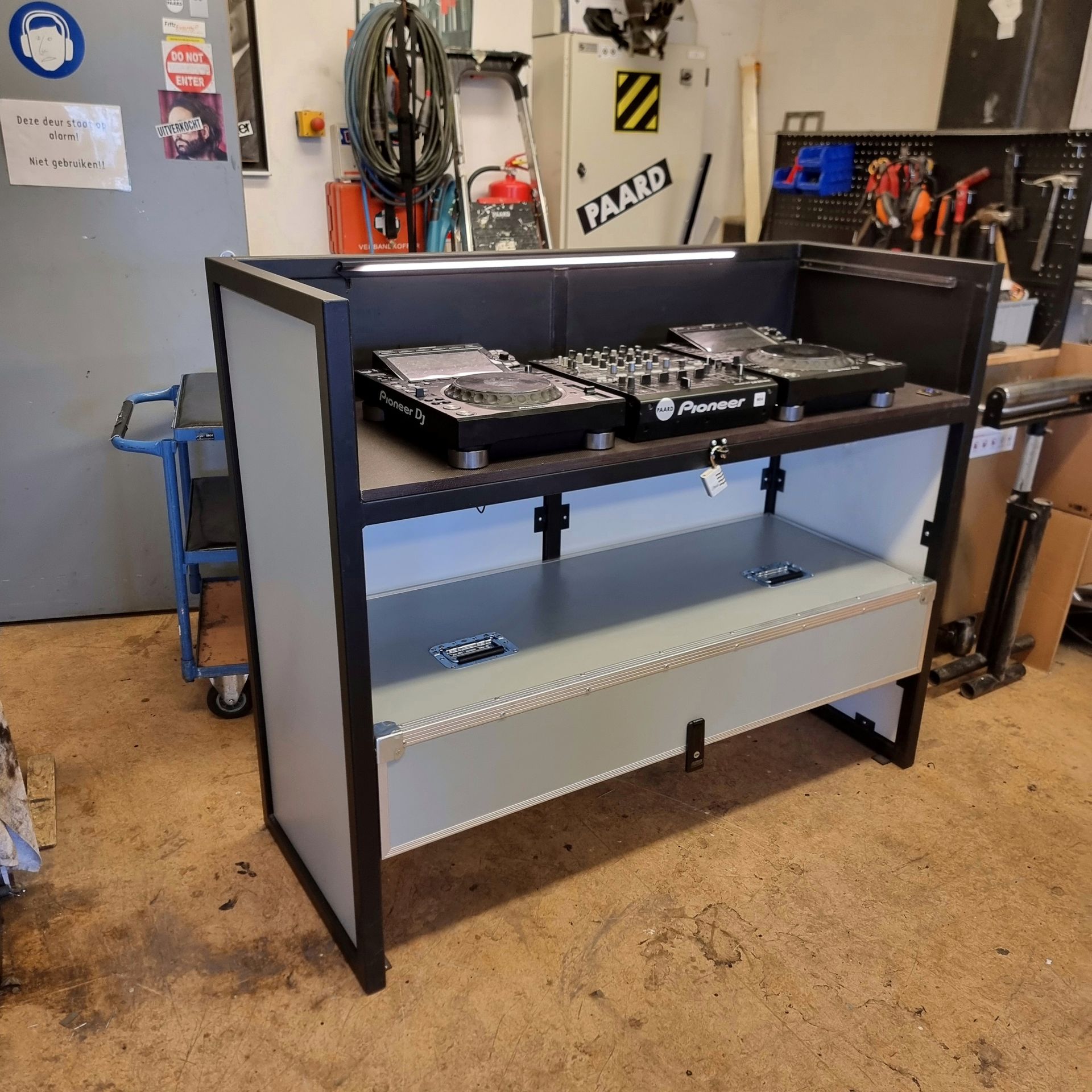 Een zwart-grijze DJ-booth met Pioneer-apparatuur staat in een werkplaats.