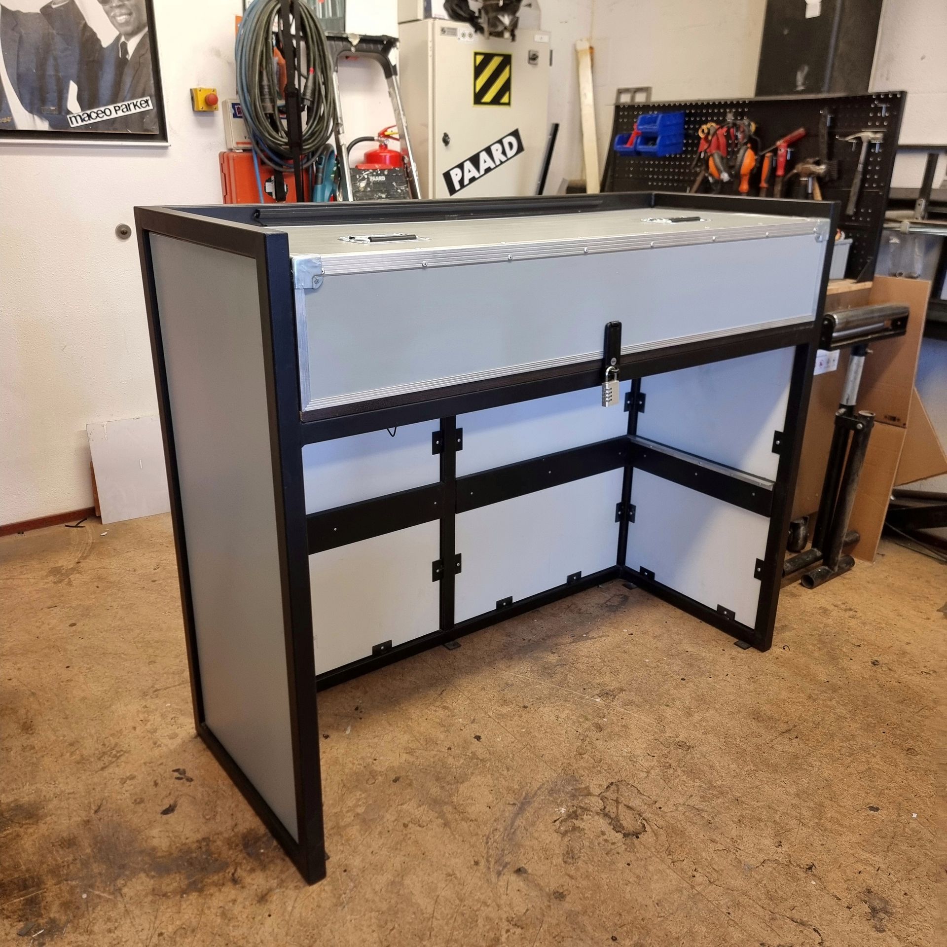 Een draagbare DJ-booth met een zwart metalen frame en lichtgekleurde panelen, opgesteld in een werkplaats of garage.