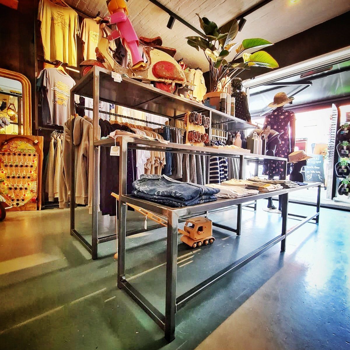 Het interieur van een kledingwinkel is voorzien van metalen displayrekken in meerdere lagen, opgehangen kledingstukken, opgevouwen spijkerbroeken en decoratieve accenten.