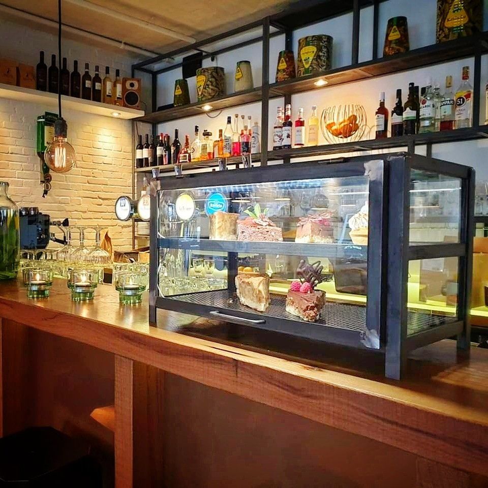 Een cafébalie is voorzien van een glazen vitrine gevuld met gebak, verlichte schappen met flessen en sfeervolle hanglampen.