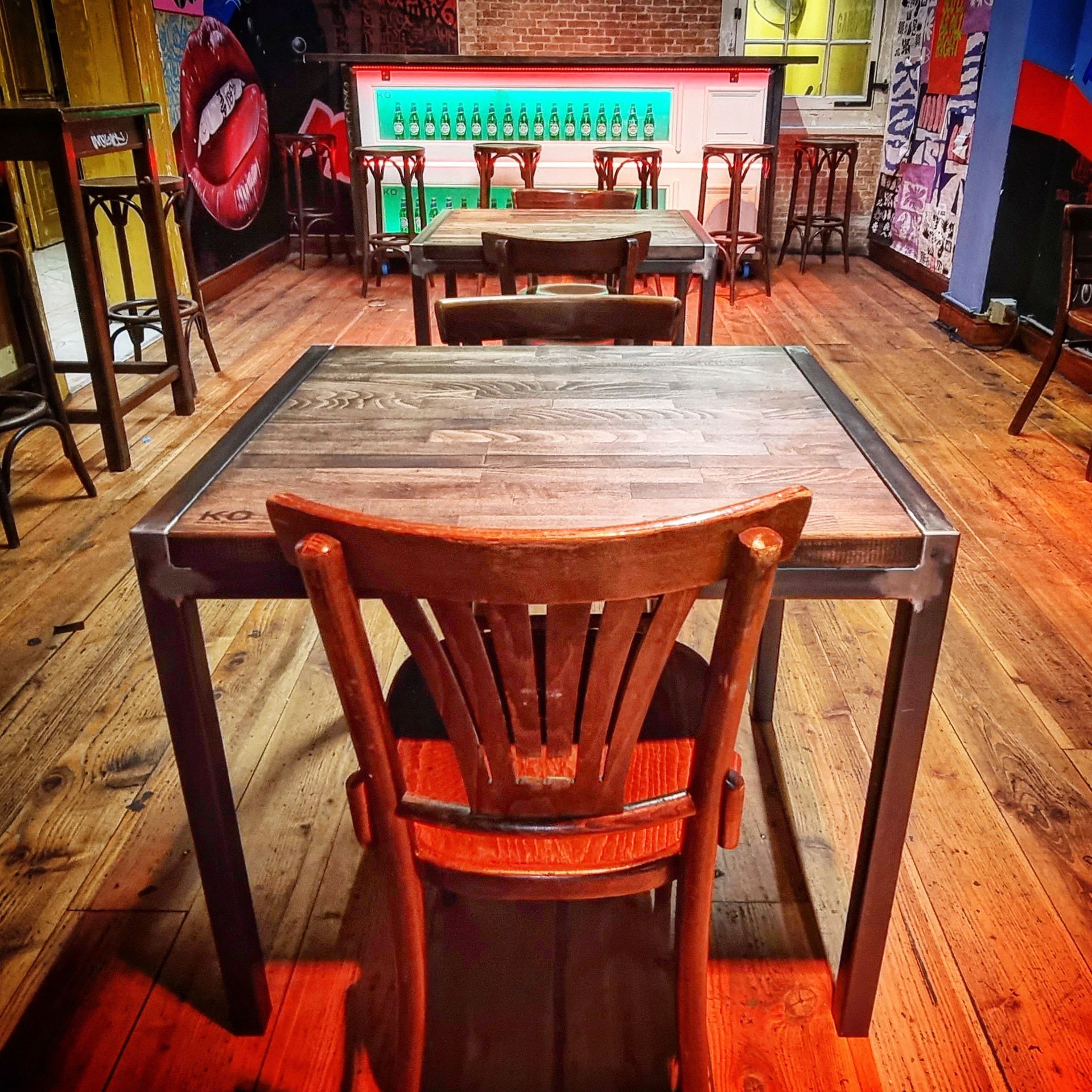Een houten tafel en stoel op de voorgrond, met daarachter meer tafels en een verlichte bar in een restaurant in rustieke stijl.