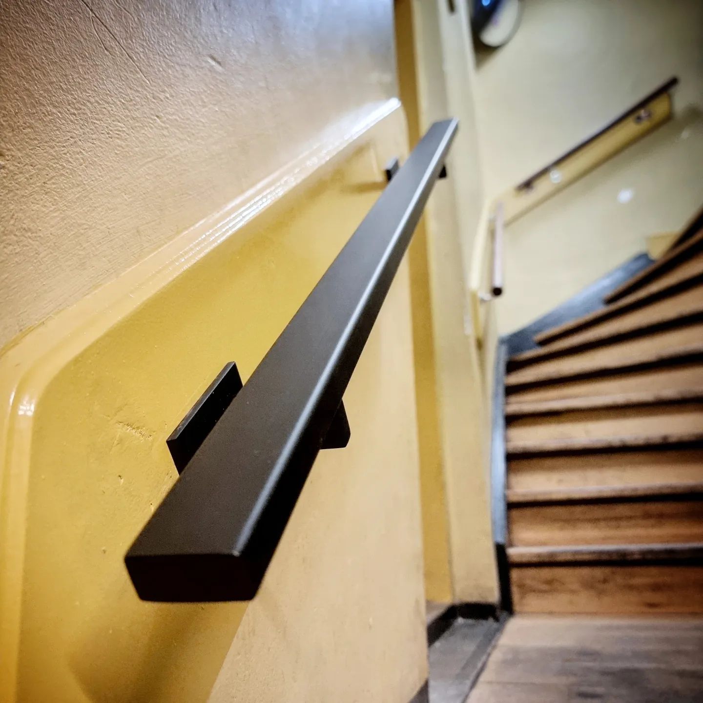 Een zwarte, rechthoekige leuning is gemonteerd aan een beige muur naast een houten trap in een schemerig verlicht trappenhuis.