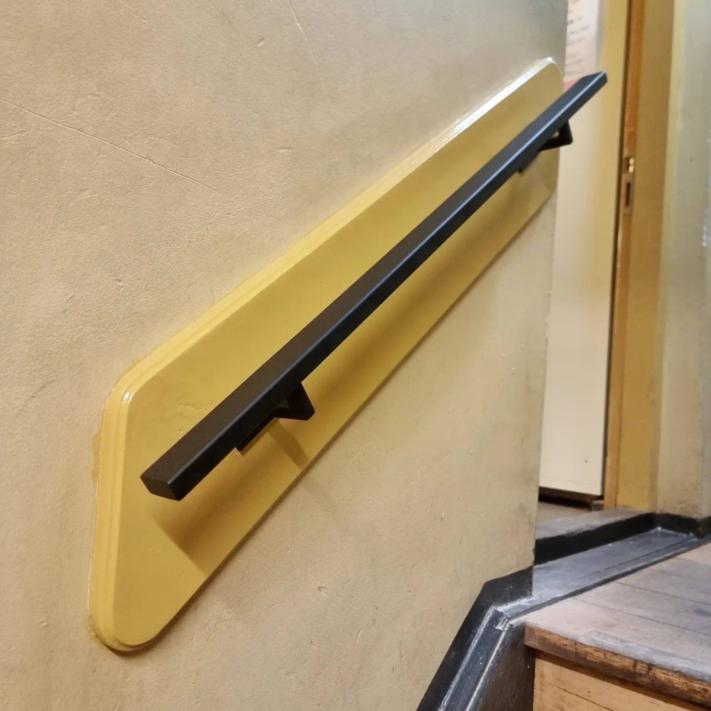 Een zwarte metalen leuning, gemonteerd op een glanzende gele muurplaat, langs een trap in een hal.