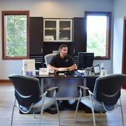 Man seated at a desk in a small office with two empty chairs and windows on either side.