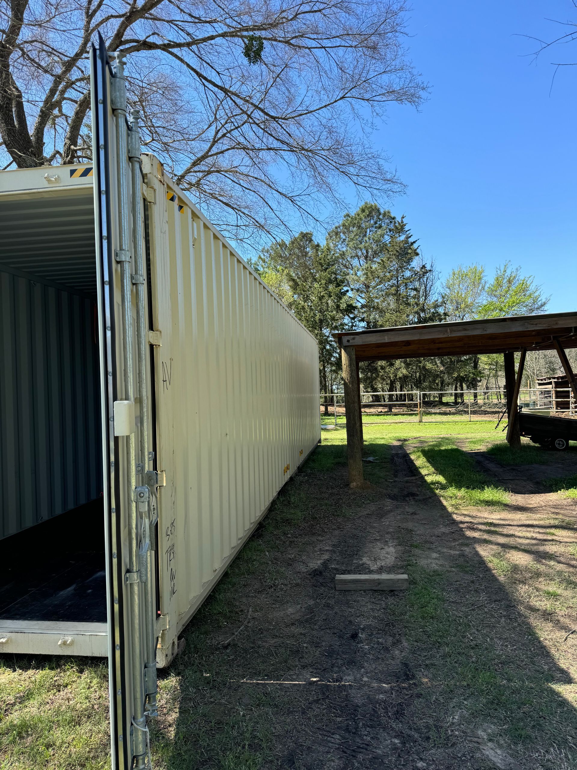Shipping container and gravel yard beside a wooden carport under a clear blue sky.