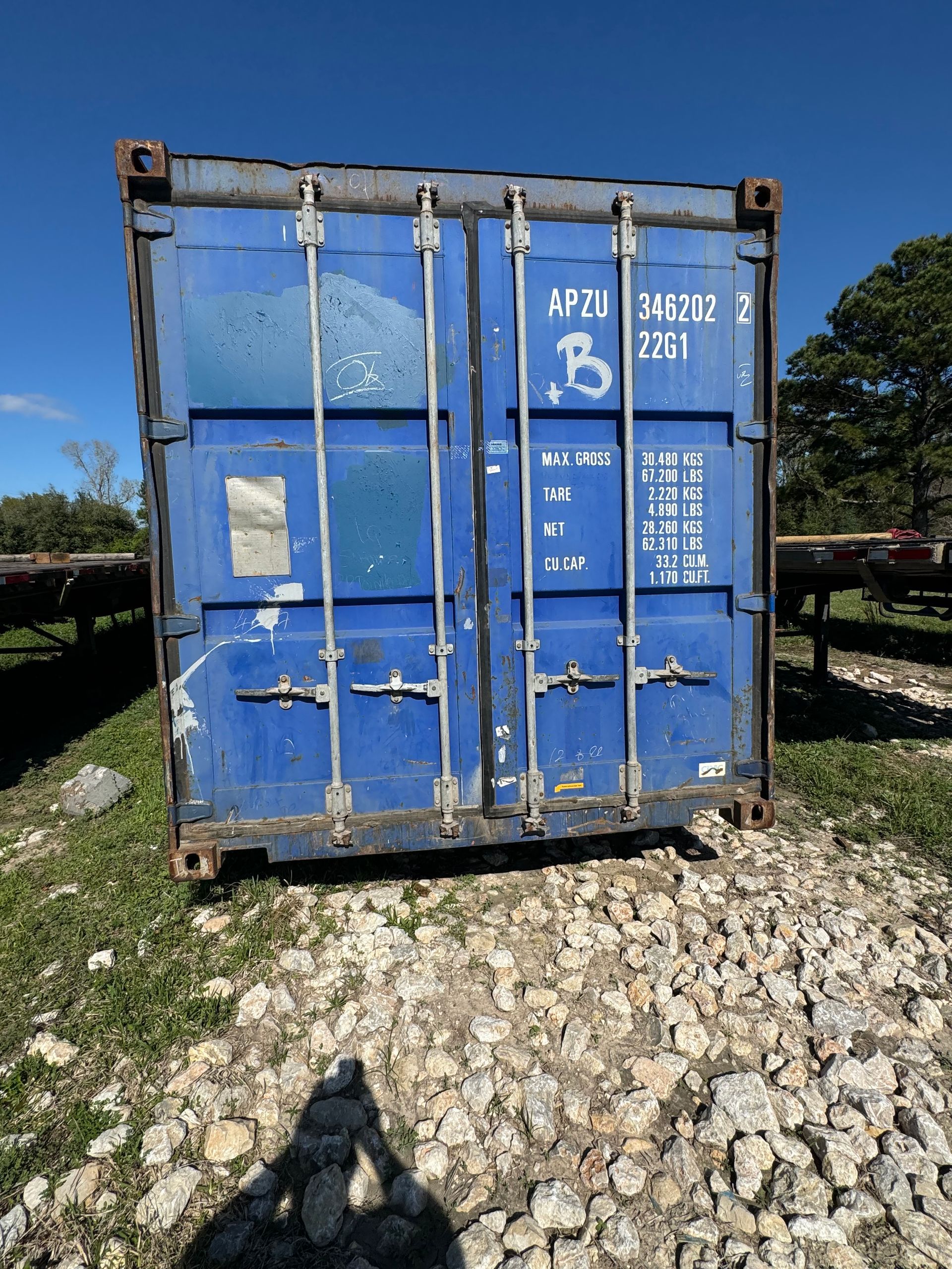 Blue shipping container on a gravel lot, with white markings and rusted corners under a sunny sky