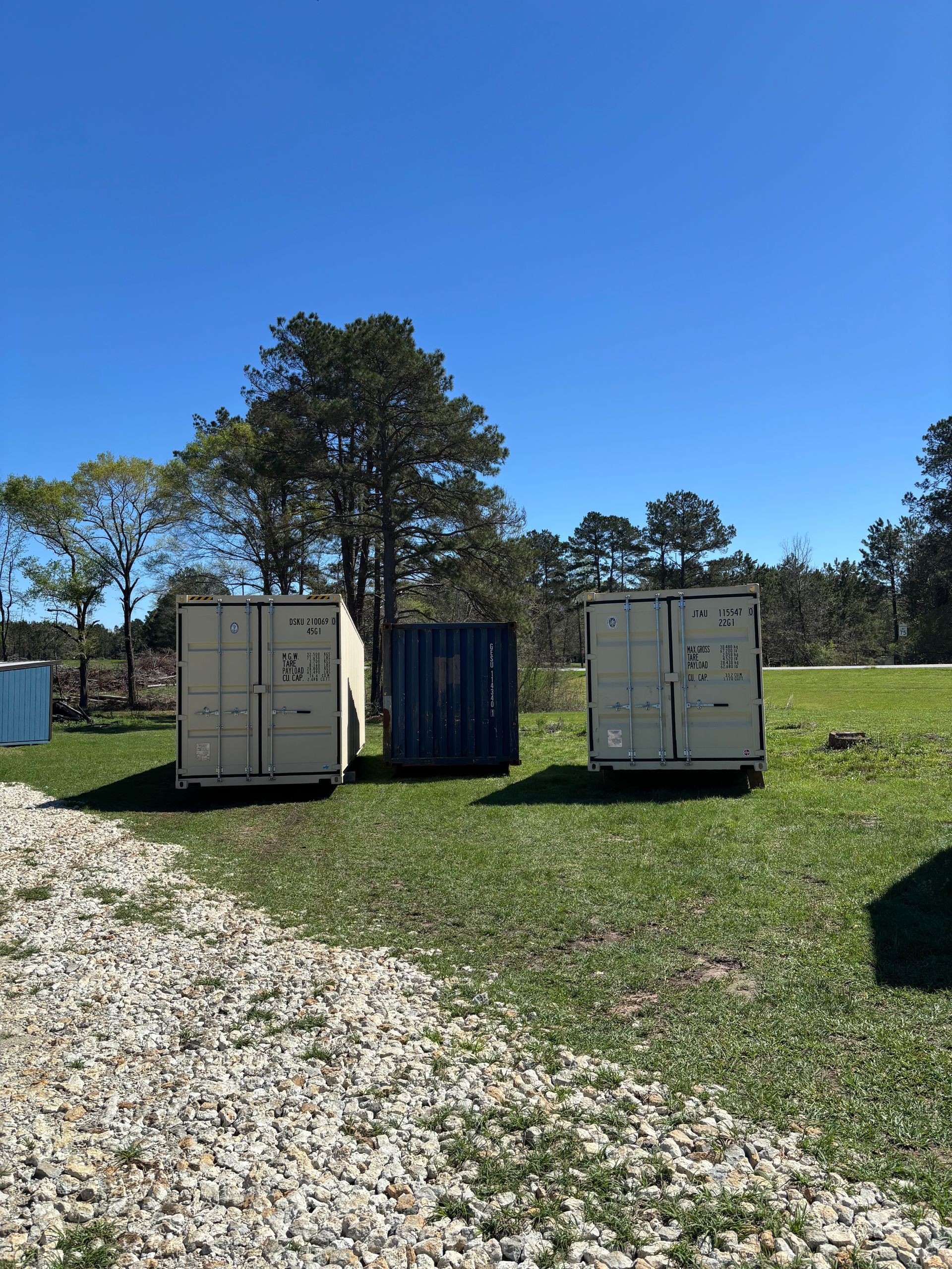 Two white shipping containers on a sunny grassy field beside a gravel path.