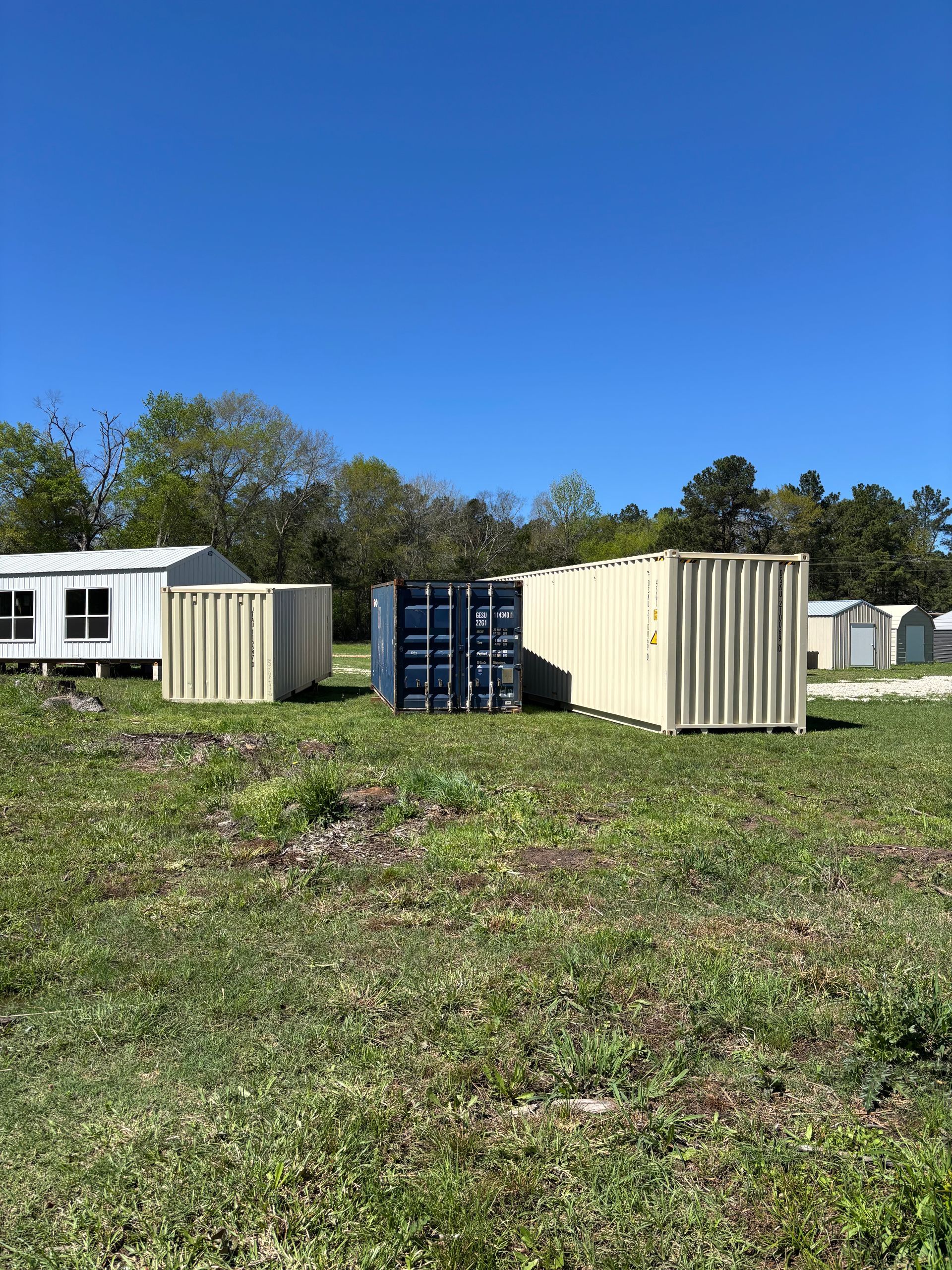 Two beige shipping containers beside a blue container on a grassy lot under a clear blue sky