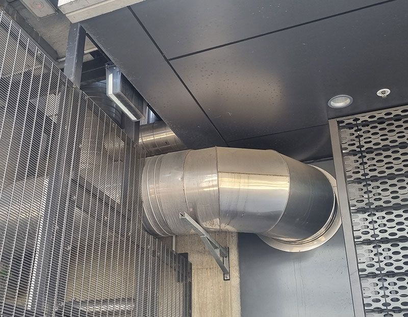 Metal ventilation ductwork installed on a building's exterior, near a metal grate and dark paneling.