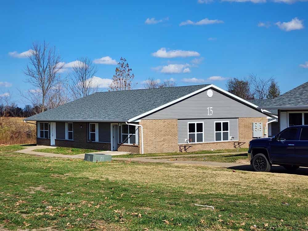 A single-story brick and gray sided building with a dark gray roof. The number 15 is visible. A blue truck is parked nearby.