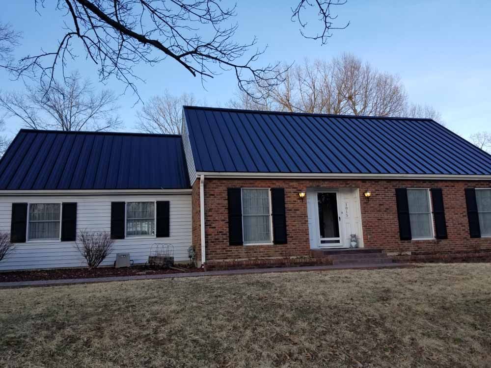 A house with a blue metal roof, white siding, and brick facade. Black shutters frame windows.