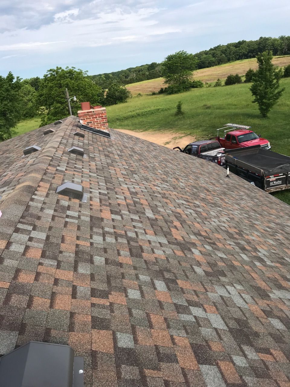 View of a roof with brown and gray shingles. A chimney is present. A truck and trailer are parked in the distance.