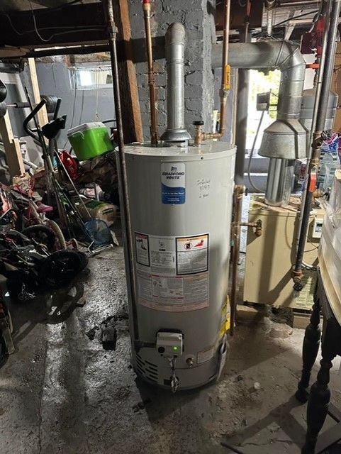 A water heater is sitting in a messy basement next to a table.