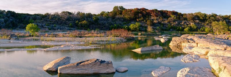 Rocky riverbank with shallow water reflecting a tree-lined hillside under warm sunset light