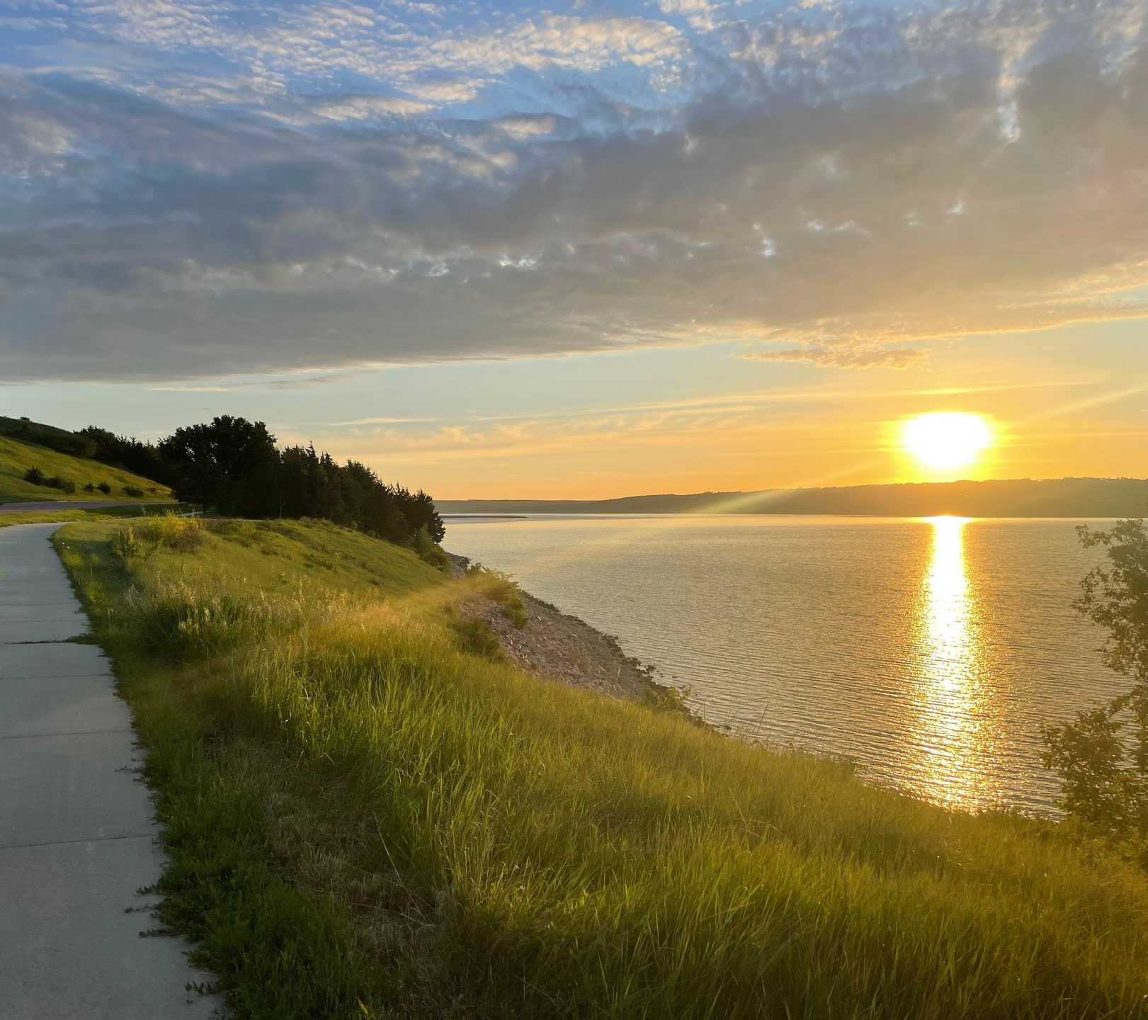 Sunset over calm water beside a grassy shoreline and paved path under a cloudy sky
