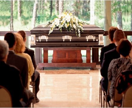 Casket with white flowers at a funeral, mourners seated in a bright chapel