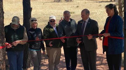 Group of people cutting a red ribbon outdoors; celebrating a trail opening.