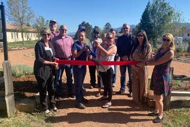 Group cutting a red ribbon on a path; sunny day, green trees in the background.