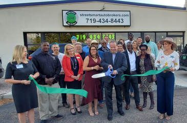 People cutting a ribbon at a business grand opening; business sign above door; light skin, many smiles.