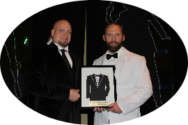 Two men in tuxedos hold award with suit silhouette. Black background.