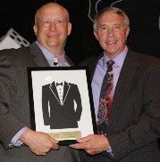Two men in suits holding a framed graphic of a tuxedo; at an event.