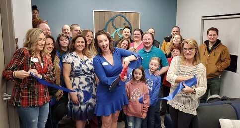 People cutting a blue ribbon at an office opening. Smiles, excited expressions, indoors.