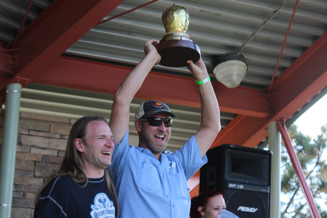 Man in blue shirt holds trophy aloft, smiling, with another man beside him. Outdoor stage setting.
