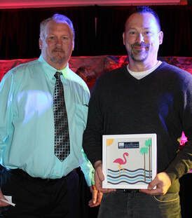 Two men posing; one holds an award with a flamingo design; they are indoors, in front of a banner.