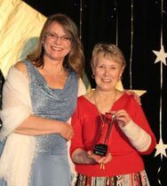 Woman in blue dress with woman in red top holding an award. Star backdrop.