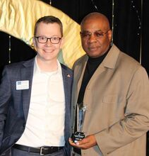 Two men smiling, one holding an award. The man on the left wears a suit and glasses. The man on the right wears a tan jacket.