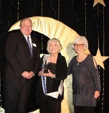 Three people on a stage: a man in a suit and two women, one holding an award. Backdrop is a crescent moon and stars.