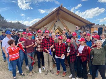 Group of people cutting a ribbon in front of a large tent. Many are wearing red hats and shirts, celebrating outside.