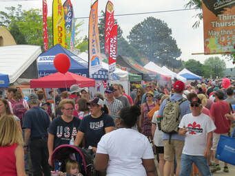 Crowd at an outdoor festival with vendor tents, flags, and balloons. People walking, wearing casual clothing.