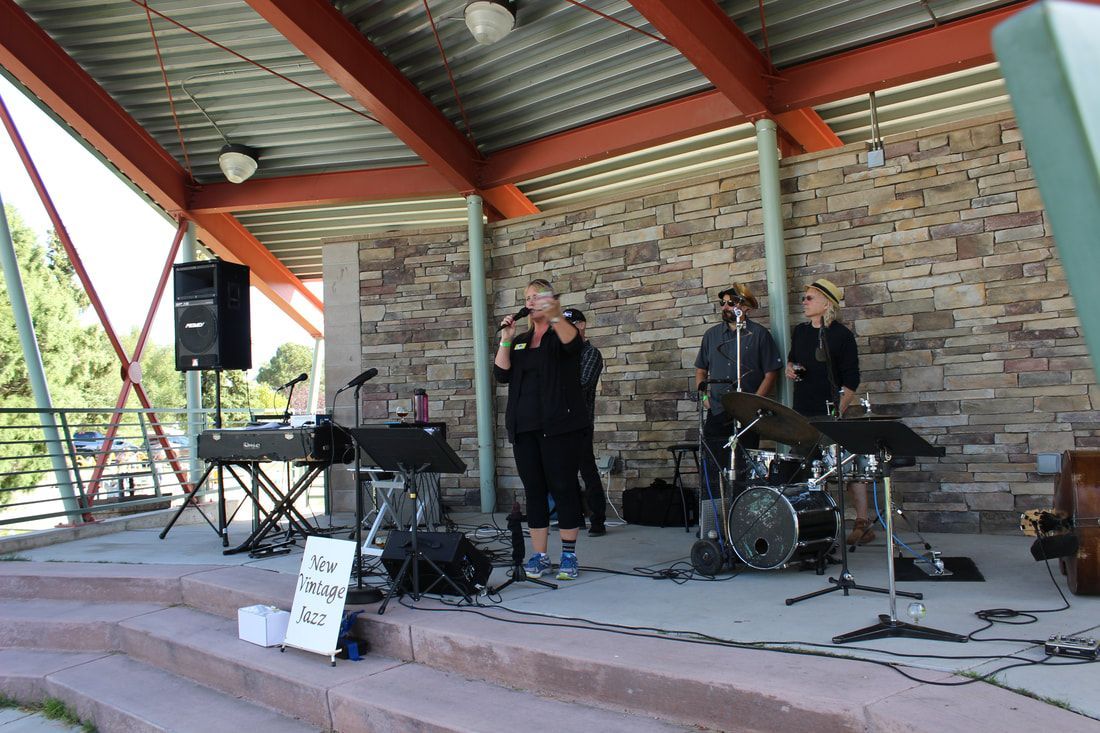 A band performs on a stage under a shelter. Musicians: three with instruments and one vocalist in front of a stone wall.